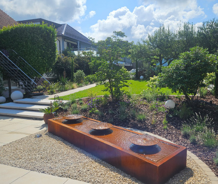 Moderne Gartenanlage mit Wasserspiel aus Cortenstahl, strukturierter Bepflanzung und klarer Wegeführung.