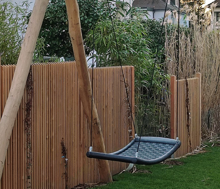 Moderner Sichtschutz aus Holzlamellen im Garten mit integrierter Schaukel und natürlicher Bepflanzung.